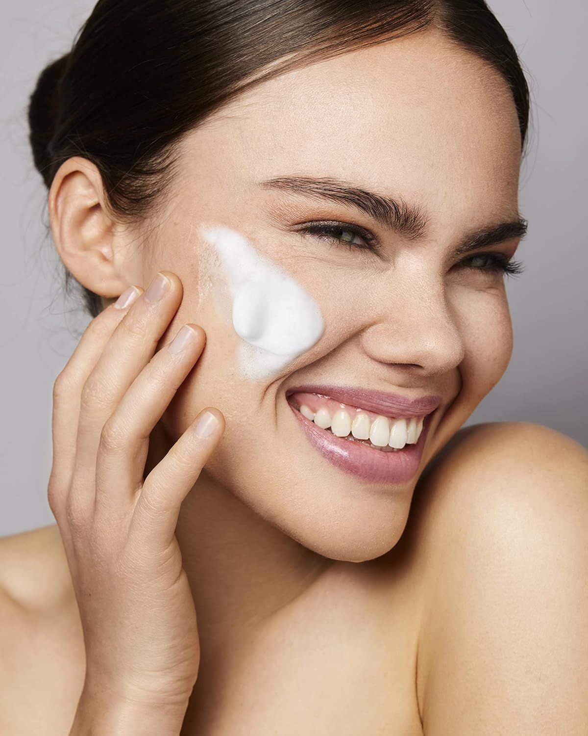 Femme avec de la mousse sur la joue, souriante. Fond: fond gris. La mousse est blanche.