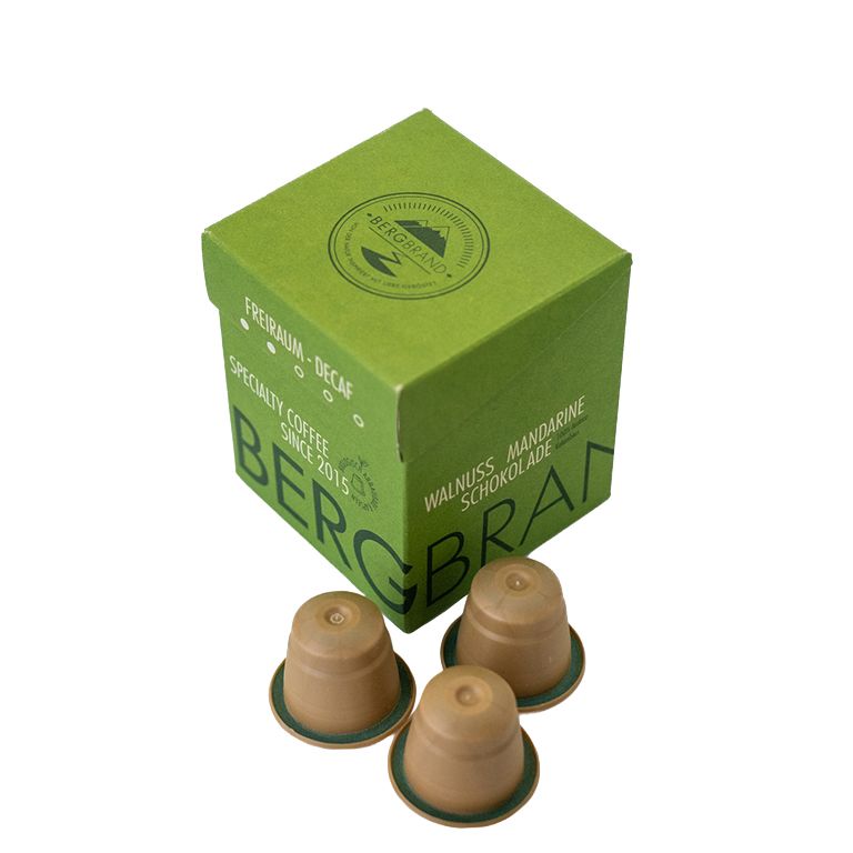 Boîte verte avec logo BERGBRAND et capsules de café. Texte : Freiraum-Decaf, Specialty Coffee, Walnuss Mandarine Schokolade.