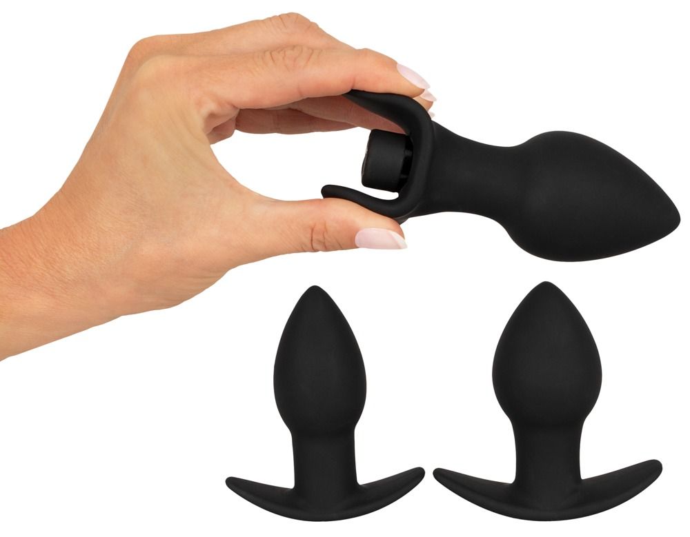 Main tenant un plug anal noir en silicone, deux autres sont posés en dessous.