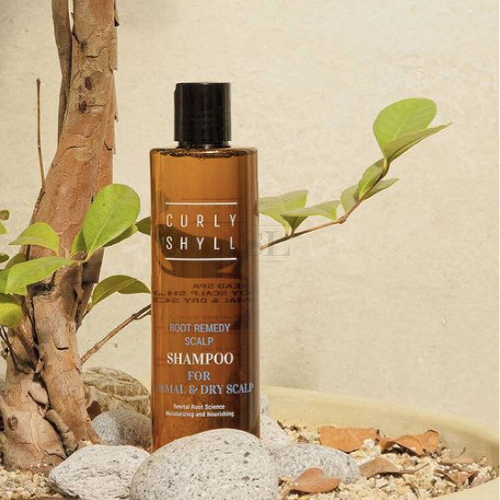 Bruine shampoo fles met zwarte dop, omringd door planten en stenen. Opschrift: CURLY SHYLL, Root Remedy Scalp Shampoo, voor normale en droge hoofdhuid.