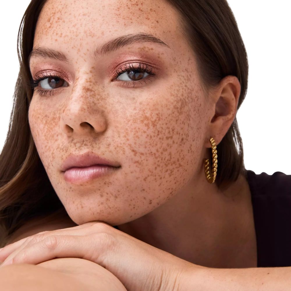 Portrait d'une femme avec des taches de rousseur. Elle porte du fard à paupières rose. Créoles dorées. Fond blanc.