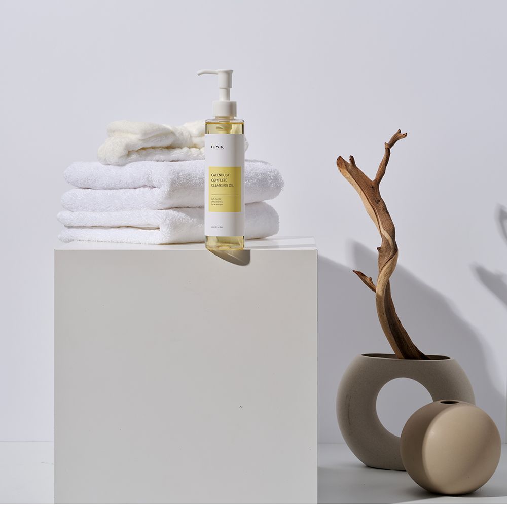 Flacon avec pompe près de serviettes et décoration. L'étiquette indique "IUNIK Calendula Complete Cleansing Oil".