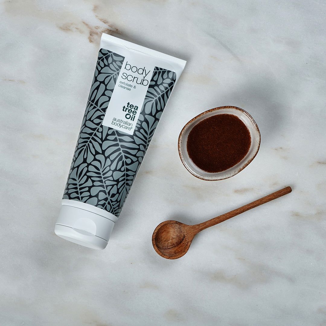 Body Scrub tube, kom en lepel. Witte tube met grijze bladeren. Tekst: Tea Tree Oil, Australian Bodycare. Bruin product.