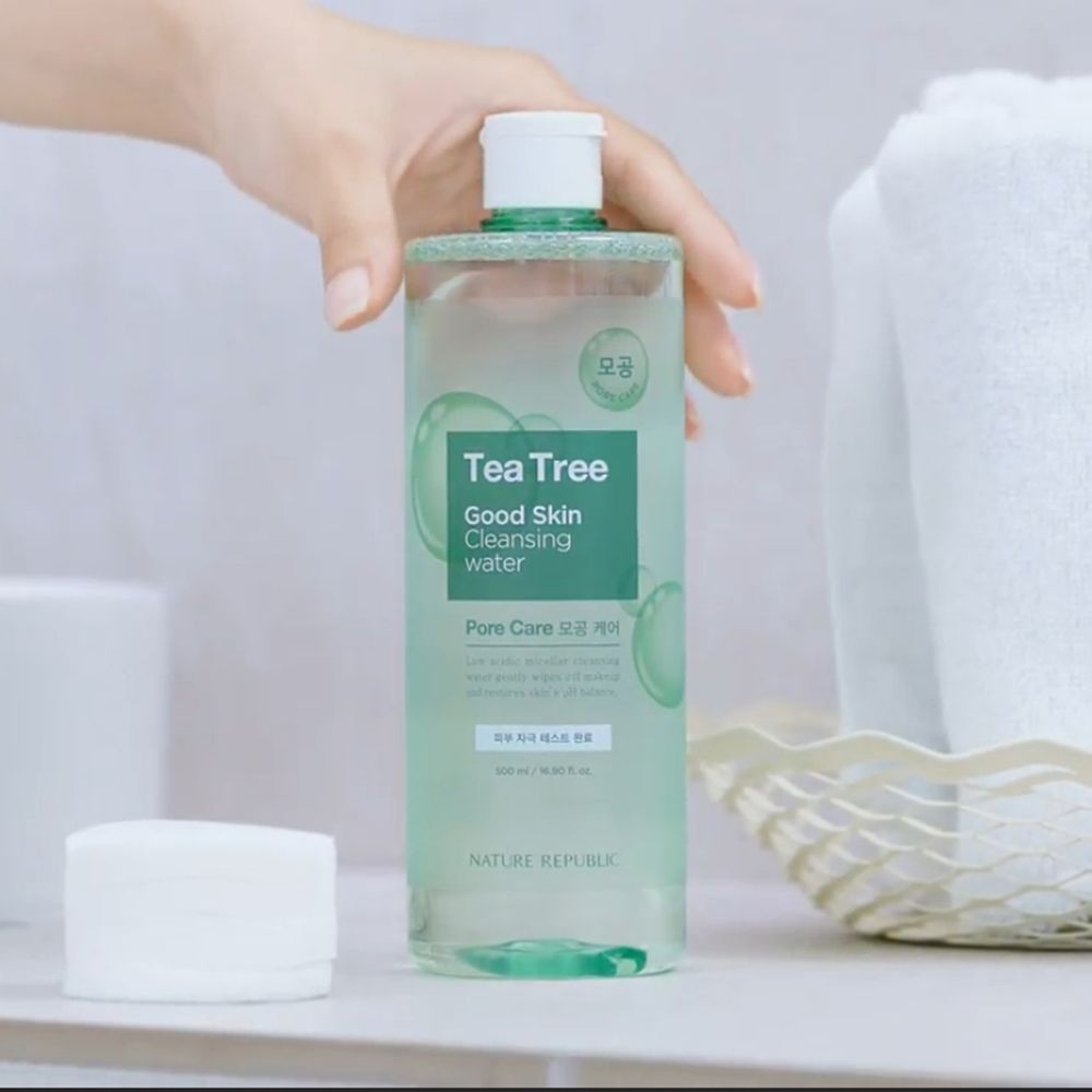 Flacon transparent avec étiquette verte. Inscription: Tea Tree Good Skin Cleansing Water. Main tenant le flacon. Disques de coton à côté.