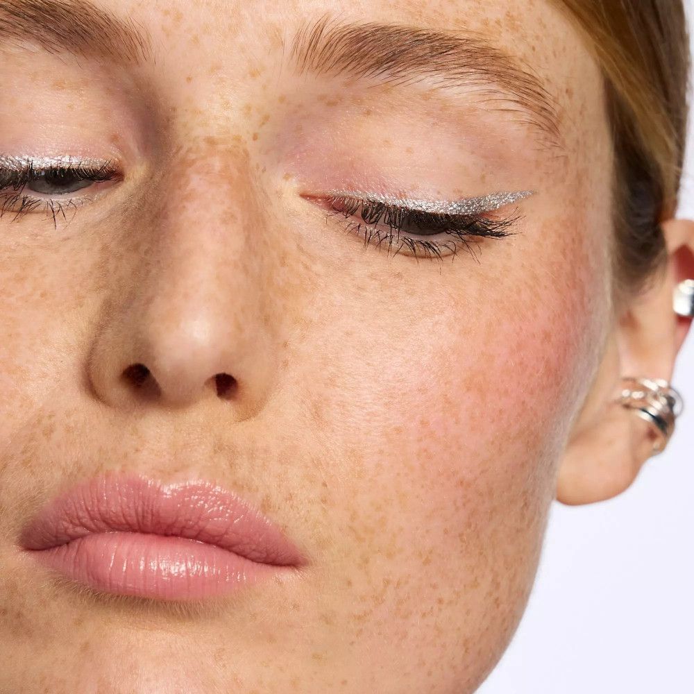 Gros plan d'un visage avec un trait d'eye-liner argenté. Yeux fermés, taches de rousseur, lèvres roses.