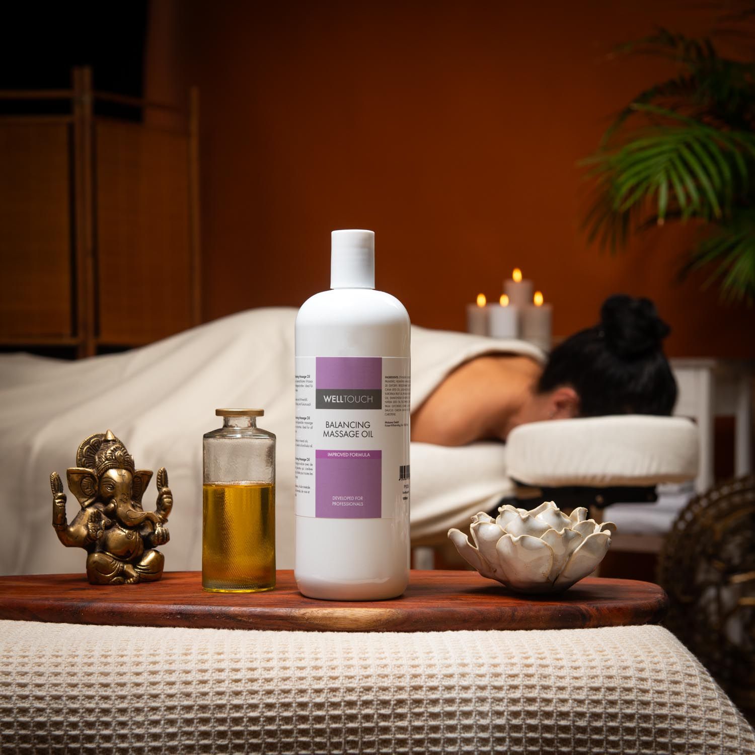Witte Welltouch massageolie, ernaast een kleine glazen fles met olie. Achtergrond: persoon op massagetafel.