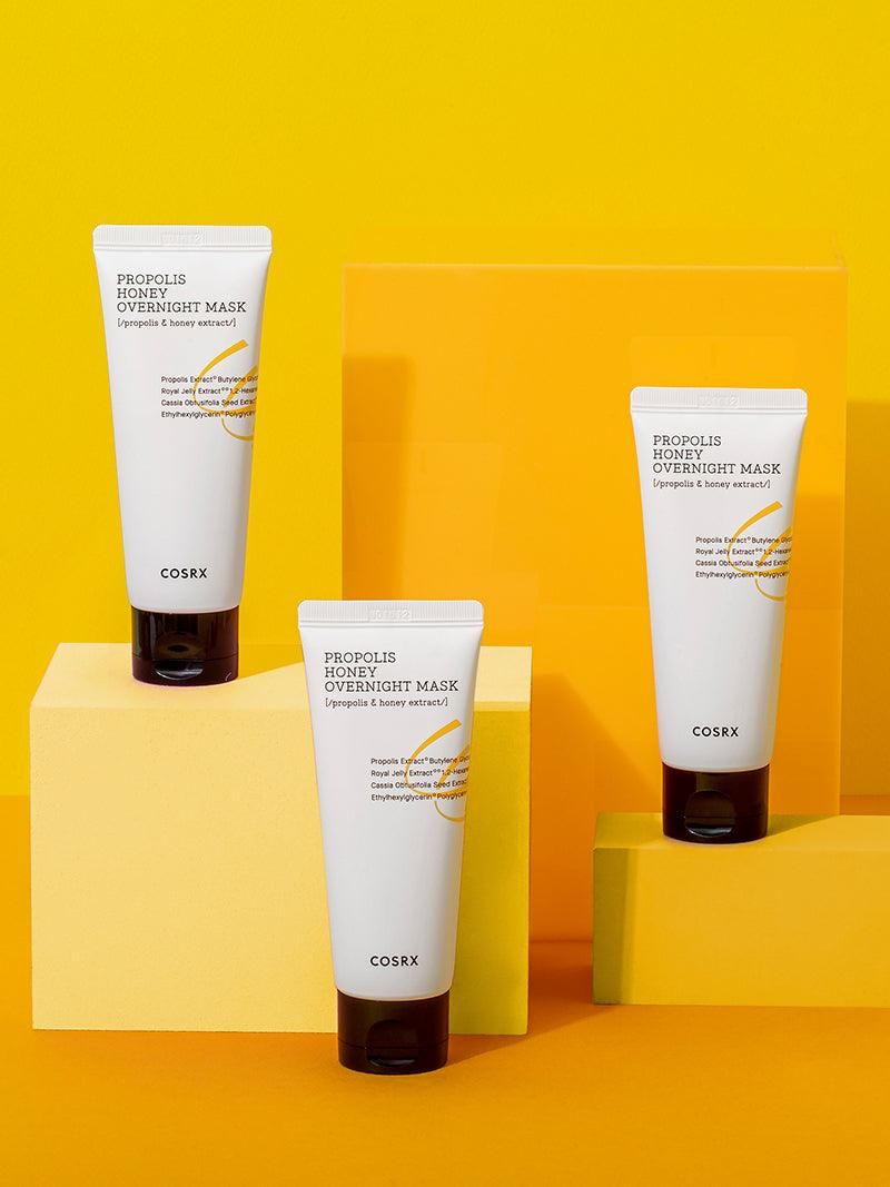Drie tubes met zwarte dop. Opschrift: Propolis Honey Overnight Mask, COSRX. Op gele achtergrond.
