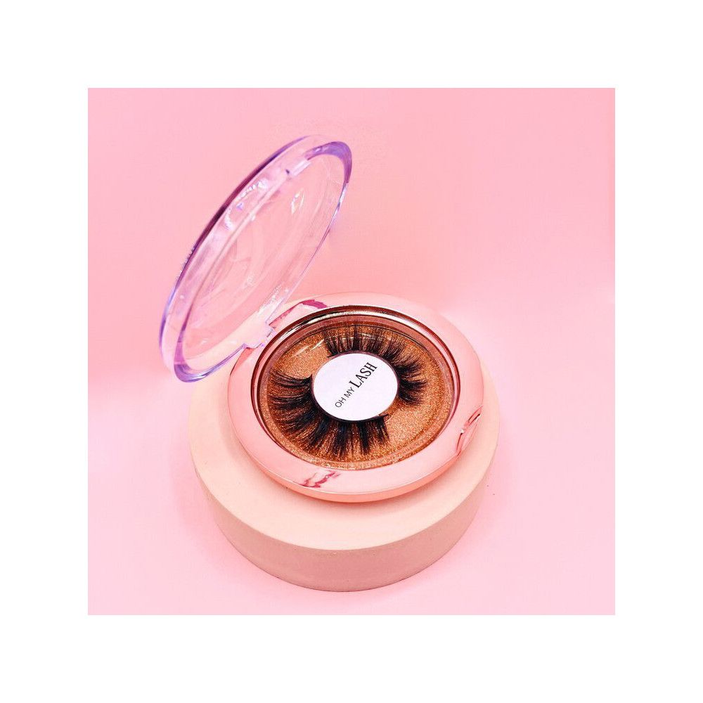 Faux cils dans un emballage rond rose. Couvercle ouvert. Inscription : OH MY LASH. Sur socle rose, fond rose.