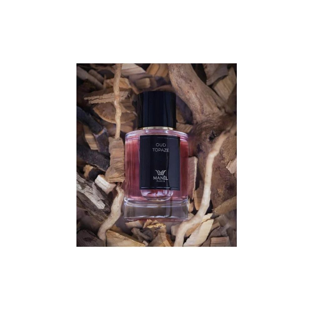 Flacon de parfum avec bouchon noir et étiquette. Inscription: Oud Topaze, Manel Paris. Posé sur des morceaux de bois.