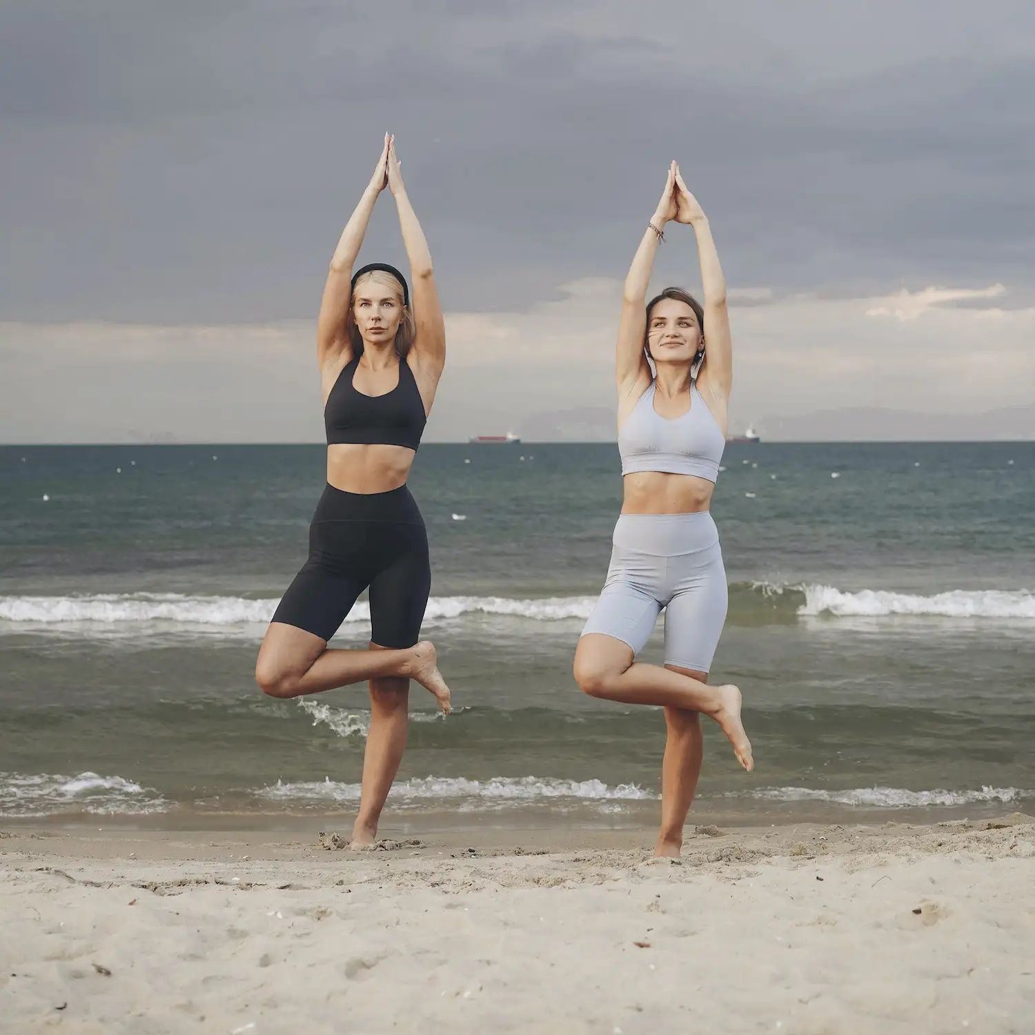 Twee vrouwen in sportkleding staan op het strand in yogahouding. Zee en lucht op de achtergrond.