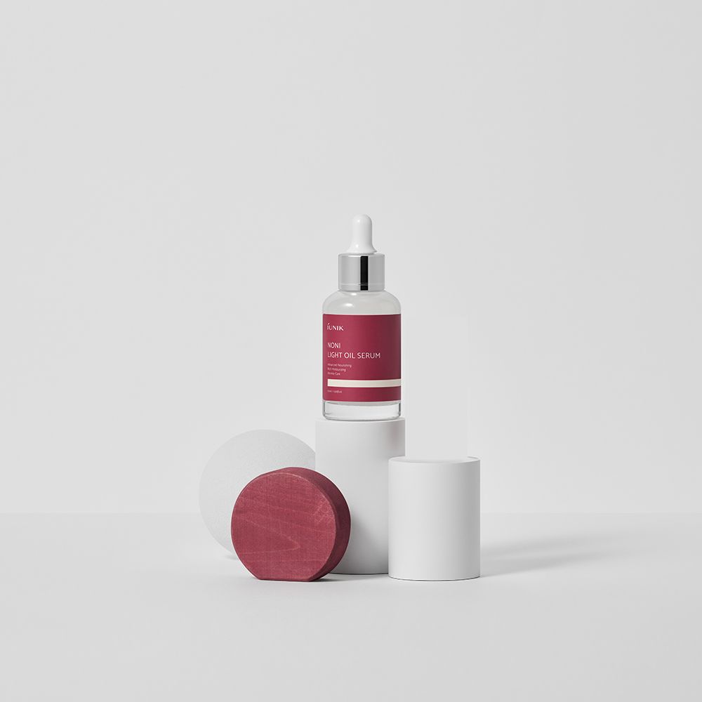 Glazen fles met rood etiket op witte kolom. Opschrift: iUNIK Noni Light Oil Serum. Met pipet.