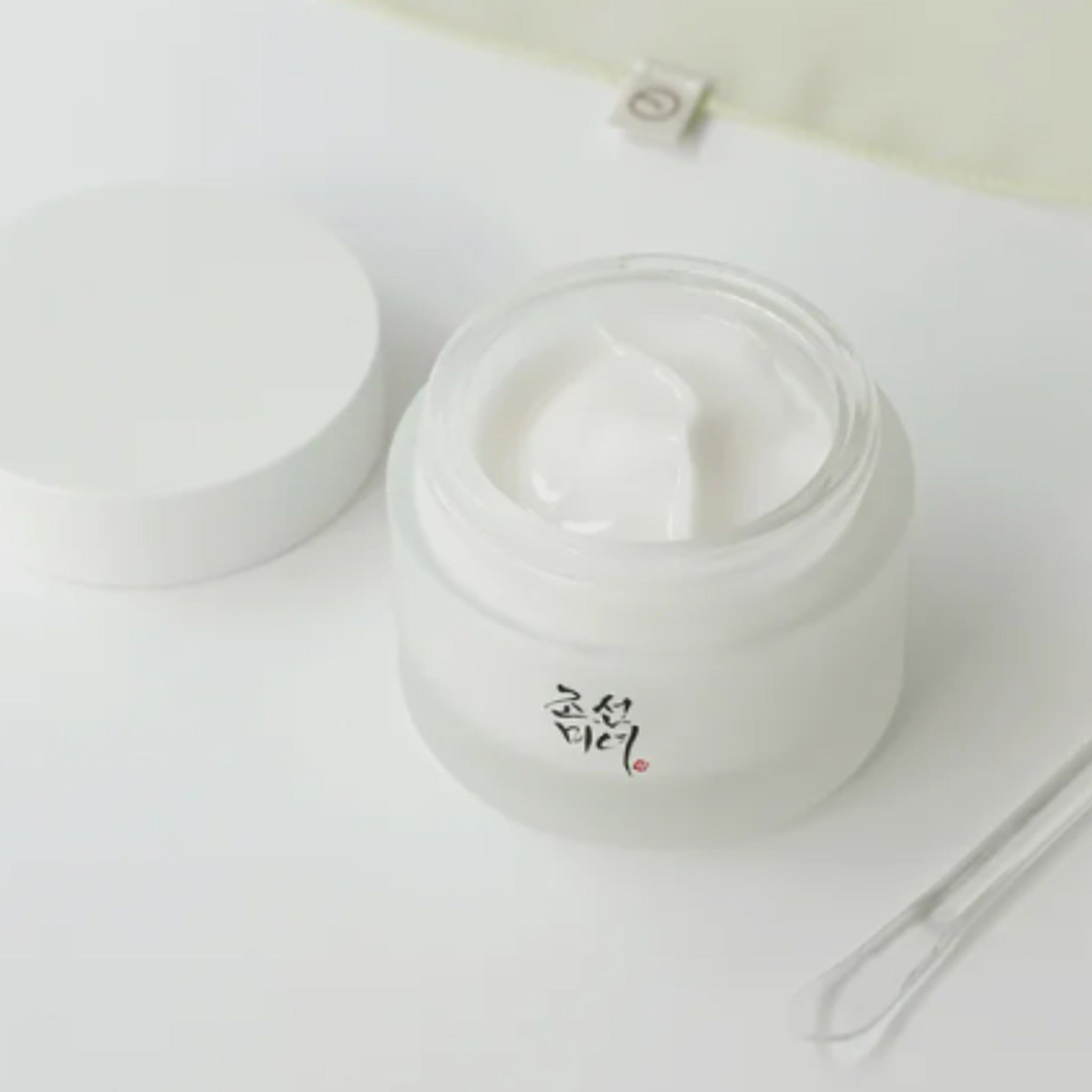 Pot de crème ouvert, couvercle à côté. Inscription coréenne. Spatule transparente.