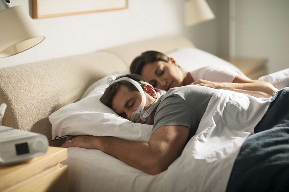 Un homme dort au lit avec un masque CPAP, une femme dort à côté. Appareil CPAP sur la table de chevet.