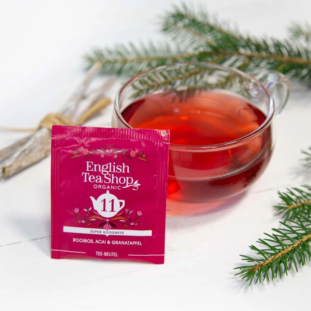 Sachet de thé avec "11 Rooibos, Acai & Grenade". Marque : English Tea Shop. Thé dans un verre. Verre avec thé rouge. Branche verte à côté.