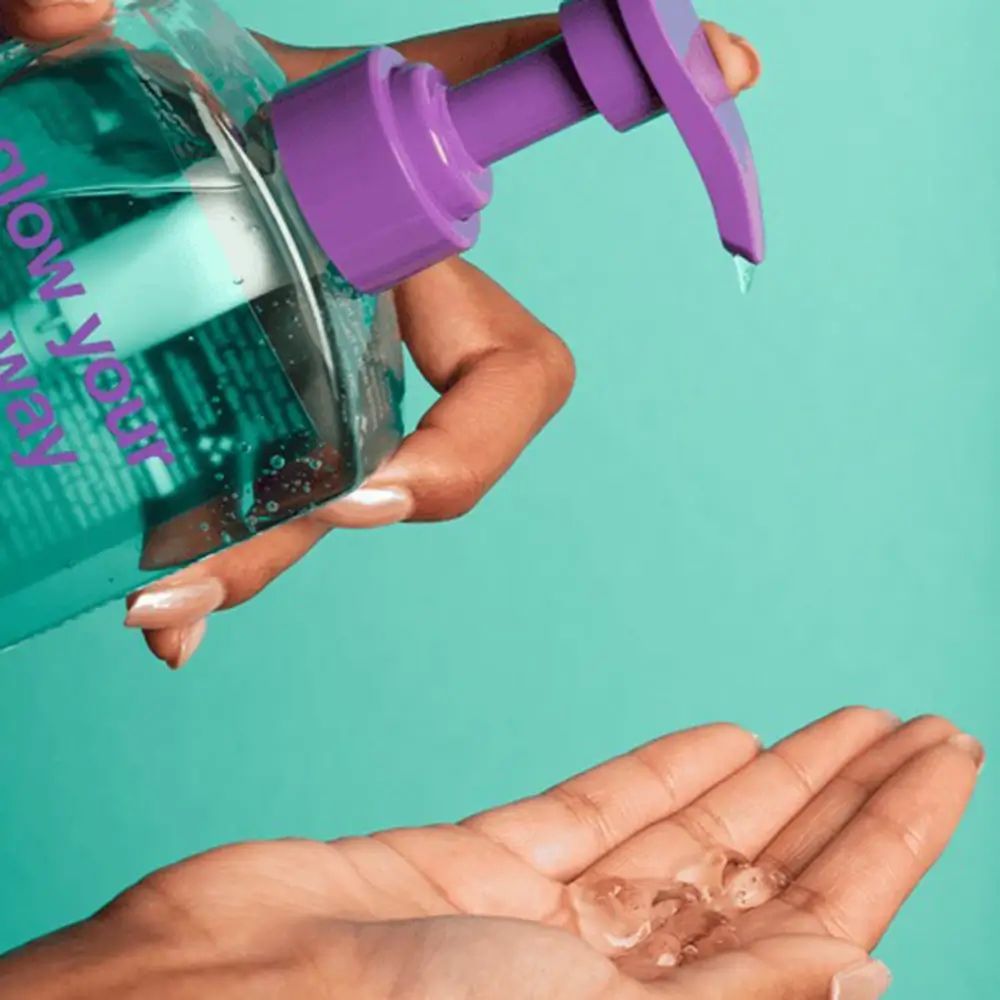 Main tenant un flacon avec une pompe violette. Gel versé dans la paume. Inscription : glow your own way.