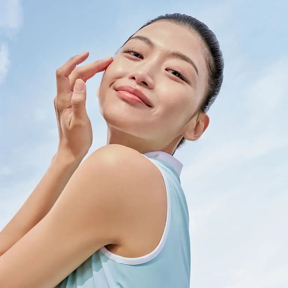 Jonge vrouw raakt zachtjes haar gezicht aan. Blauwe lucht op de achtergrond. Ze glimlacht.