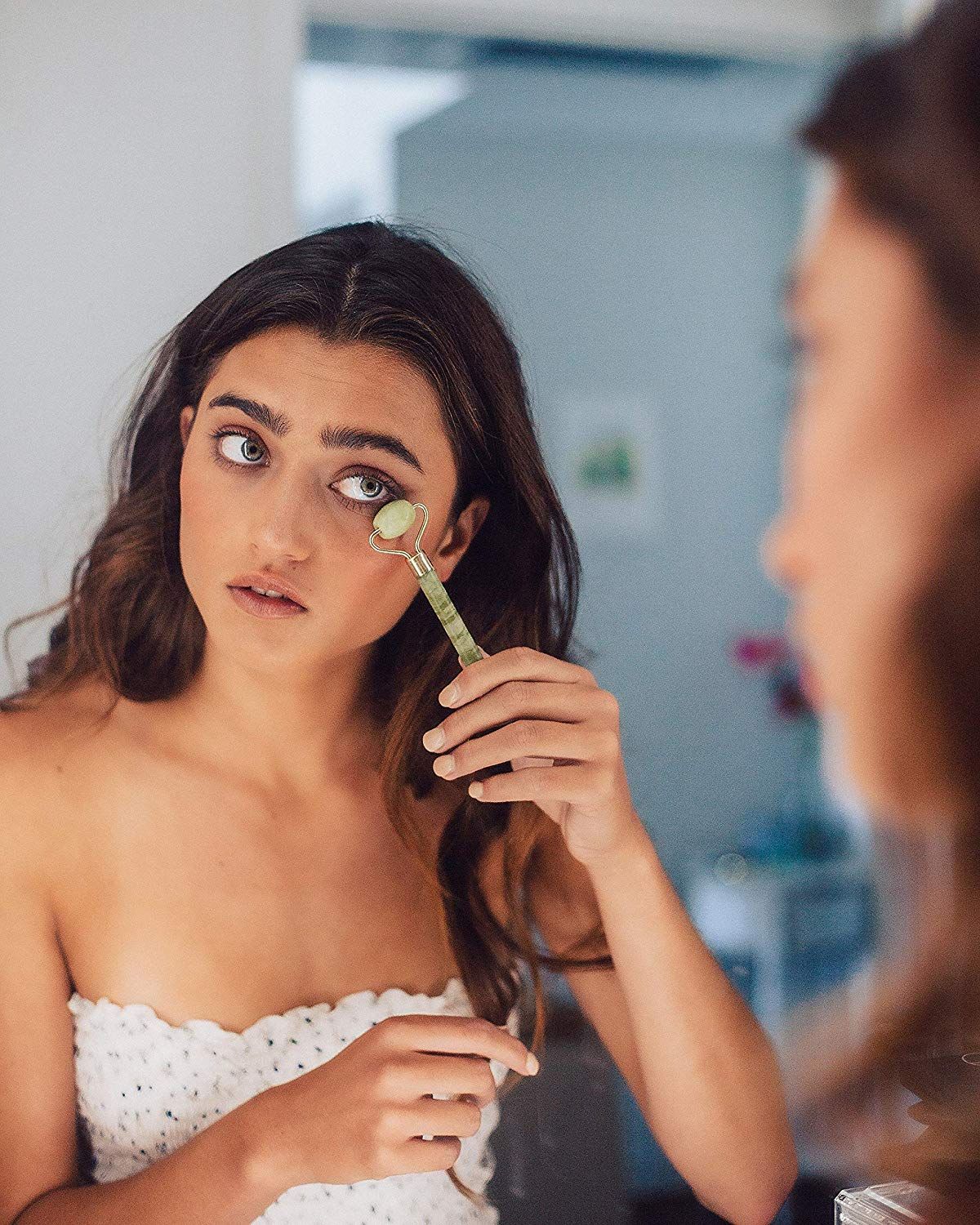 Jeune femme aux yeux bleus utilise un rouleau facial en jade vert sous son œil.