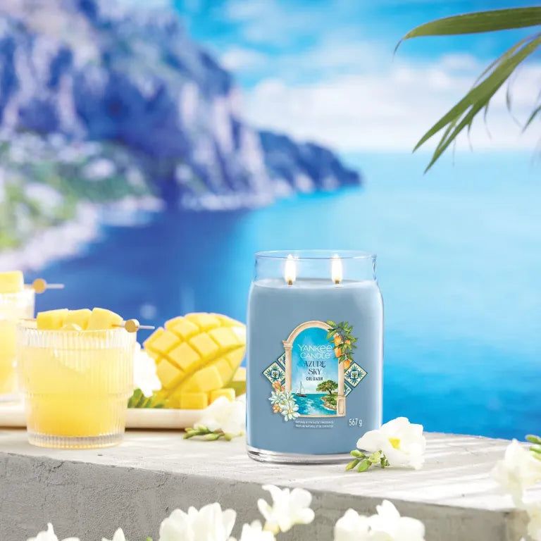 Blauwe Yankee Candle Azur Sky Signature Grand Pot met twee lonten. Opschrift op etiket. Achtergrond: zee en cocktails.