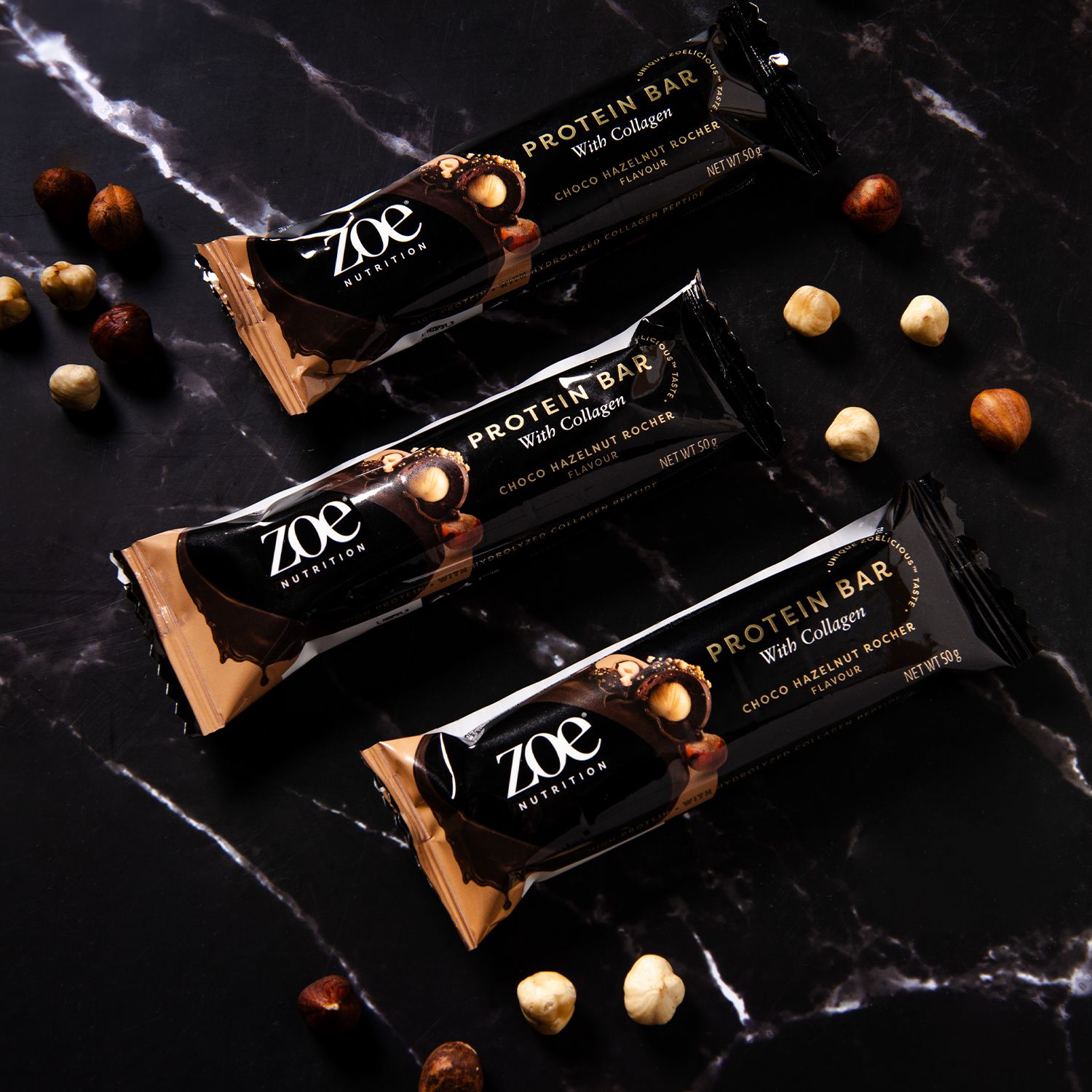 Drie eiwitrepen met collageen op zwarte marmeren achtergrond. Logo en productnaam zichtbaar. Hazelnoten verspreid.