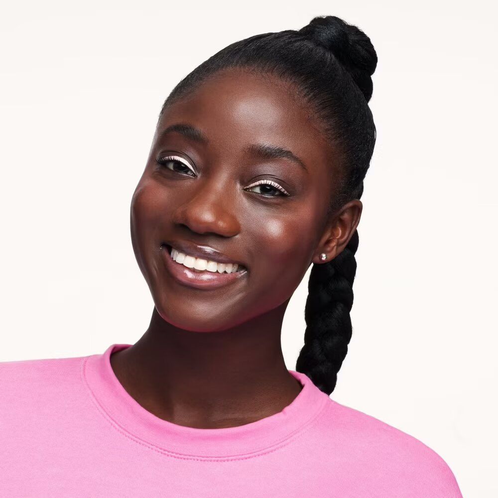 Portrait d'une femme à la peau foncée avec une tresse. Elle porte un sweat-shirt rose et sourit. Eyeliner blanc.