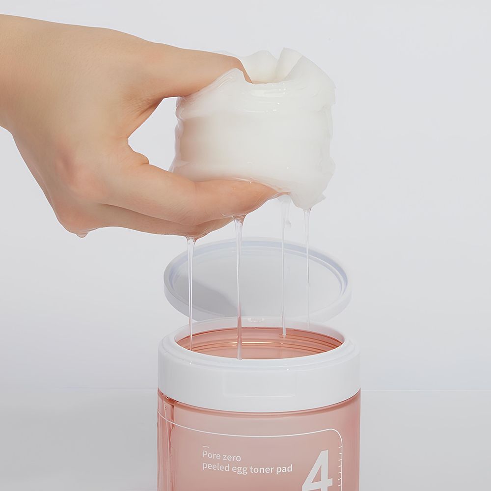 Hand houdt een gedrenkte pad boven een open pot. Vloeistof druppelt. Roze pot met witte deksel.