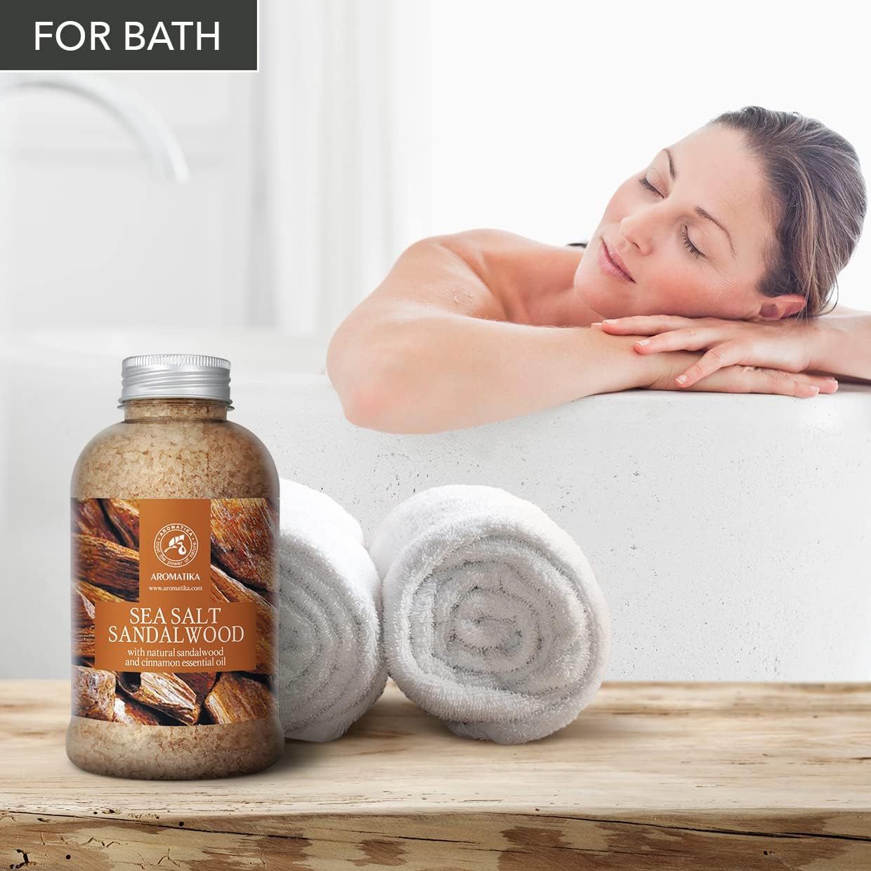 Femme se détendant dans une baignoire à côté de sel de bain AROMATIKA bois de santal et serviettes.