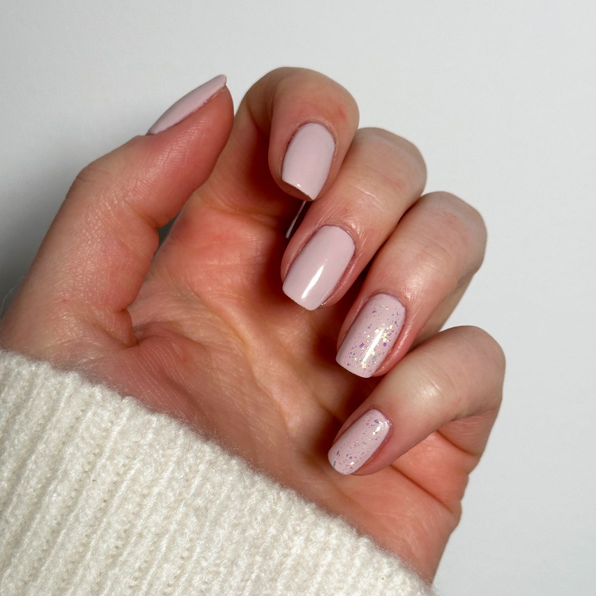 Main avec ongles peints en rose clair. Un ongle avec des paillettes. La personne porte un pull blanc.
