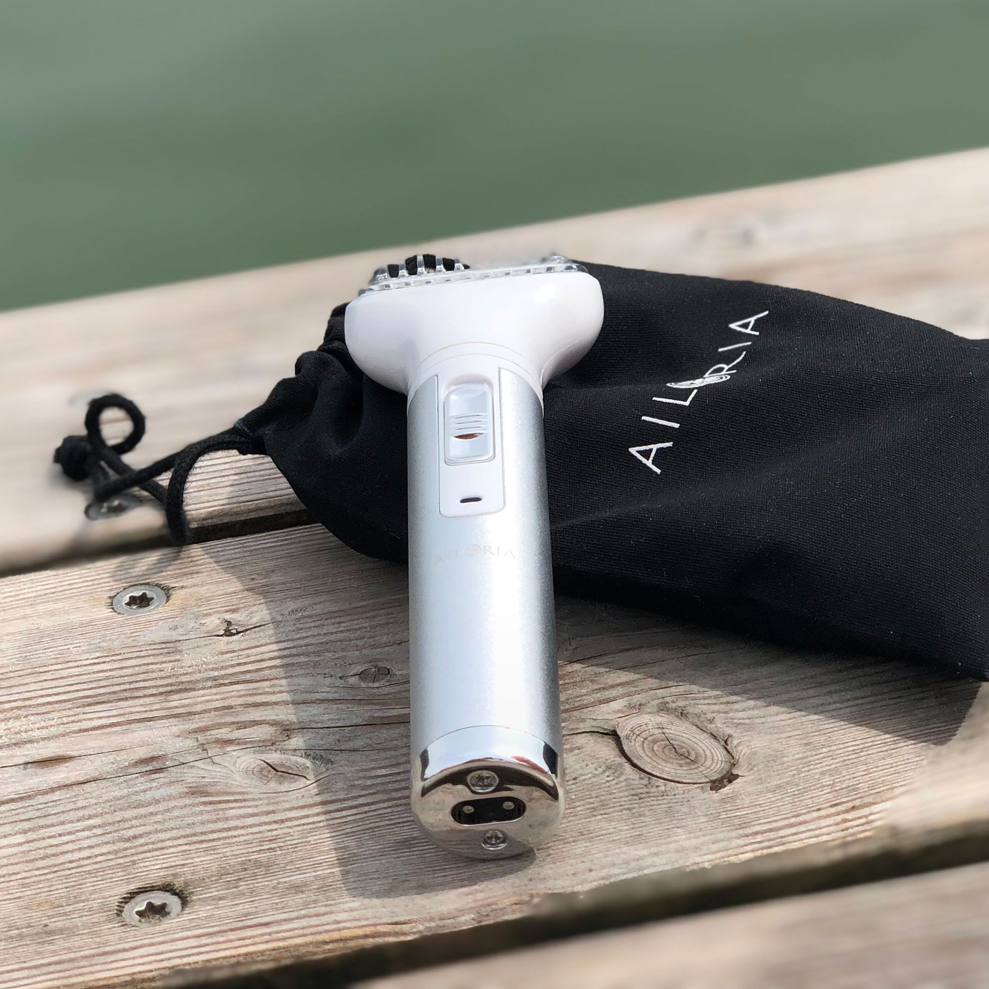 Rasoir argenté et blanc avec logo AILORIA sur pochette noire. Posé sur du bois. Rasoir avec accessoire de coupe.