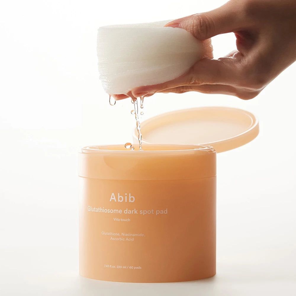 Beige pot met deksel, pads en vloeistof. Hand houdt pads boven de pot. Opschrift: Abib Glutathiosome dark spot pad.