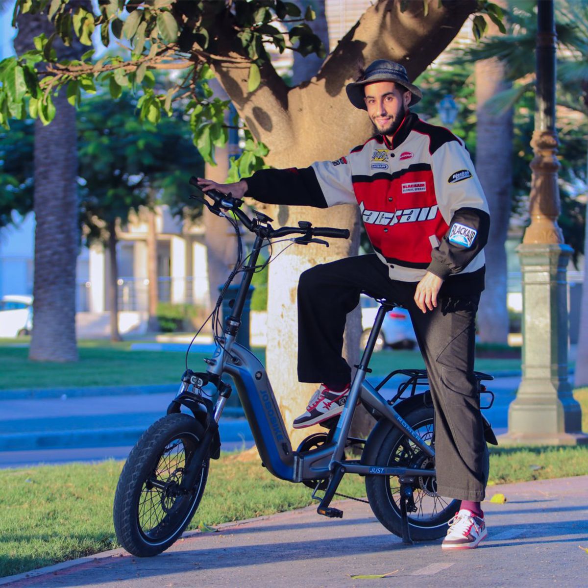 Homme assis sur un vélo électrique JOBOBIKE Romer X bleu. Il porte une veste et un chapeau. Le vélo est sur un chemin.