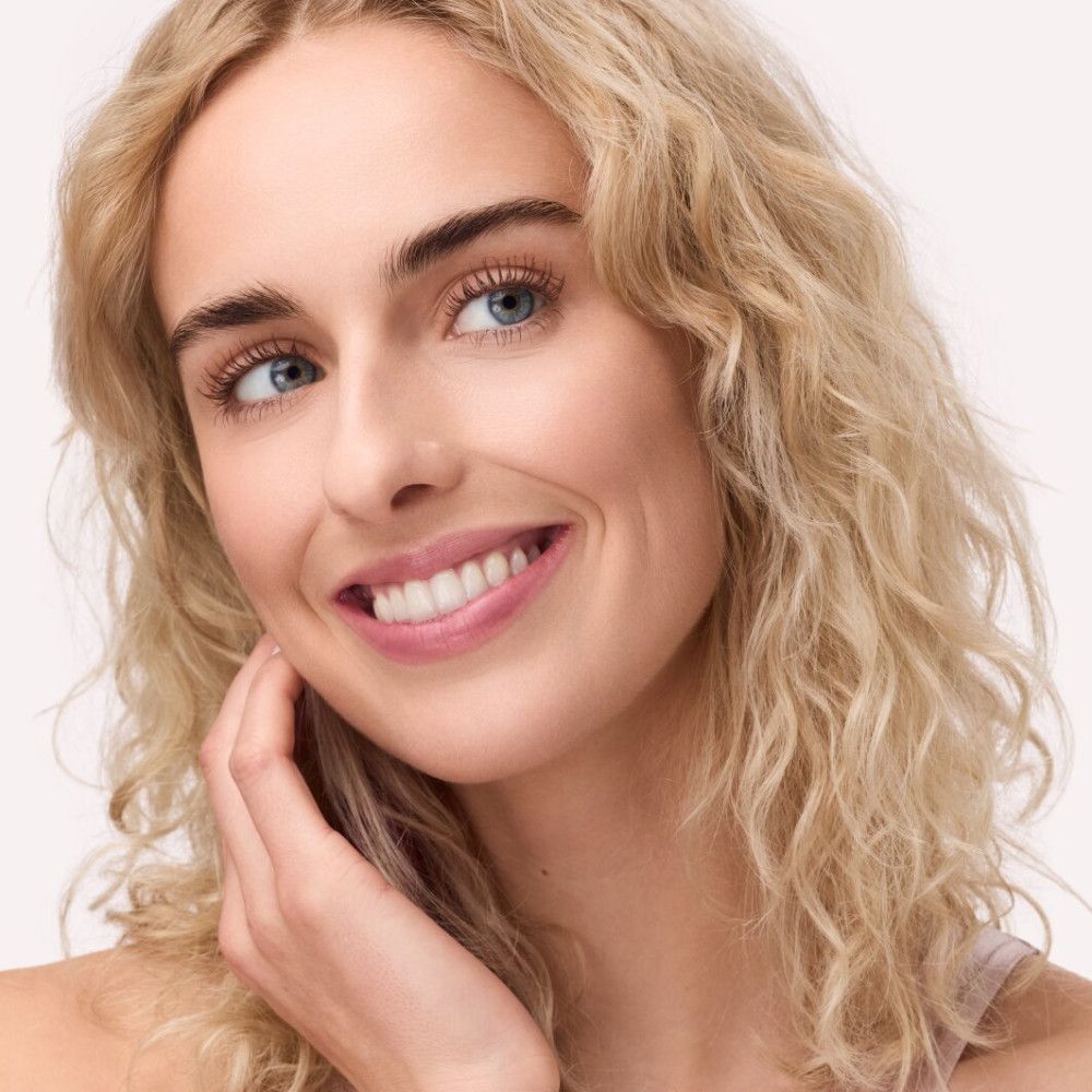 Portrait d'une femme aux cheveux blonds bouclés et aux yeux bleus. Elle sourit et touche sa joue avec sa main.