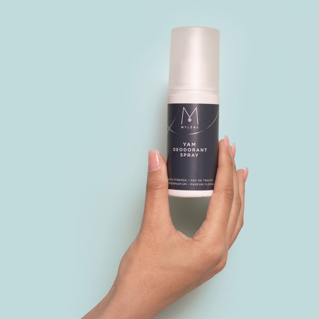 Main tenant une bouteille de déodorant Mylène Yam sur fond bleu clair.