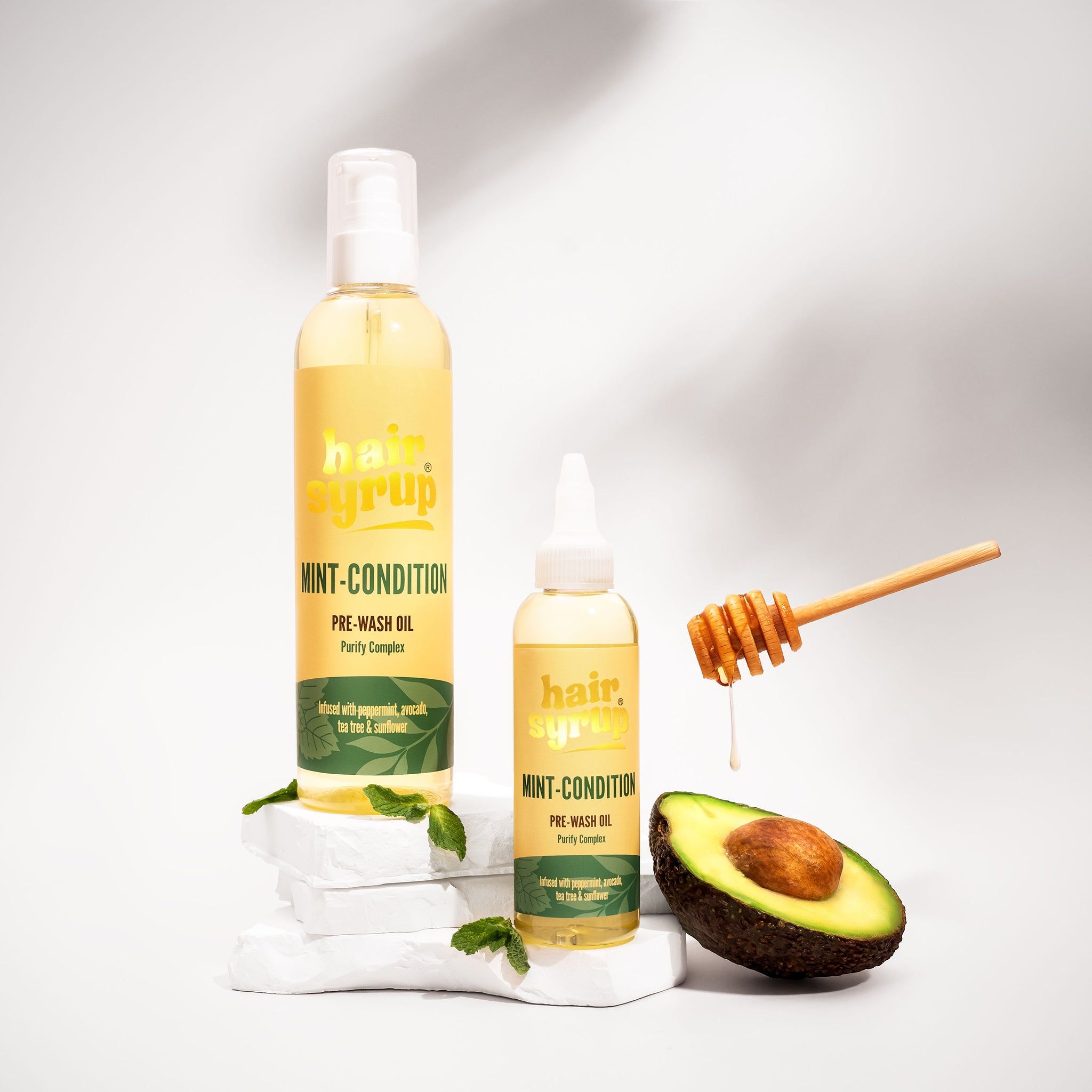 Twee flessen "Hair Syrup Mint Condition" naast avocado en honing. Op witte stenen.
