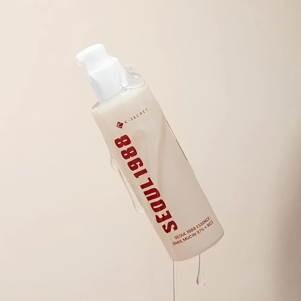 Beige fles met witte pomp. Rode tekst: SEOUL 1988. Vloeistof druppelt. Tekst: K-SECRET, SEOUL 1988 ESSENCE, SNAIL MUCIN 97% + RICE.