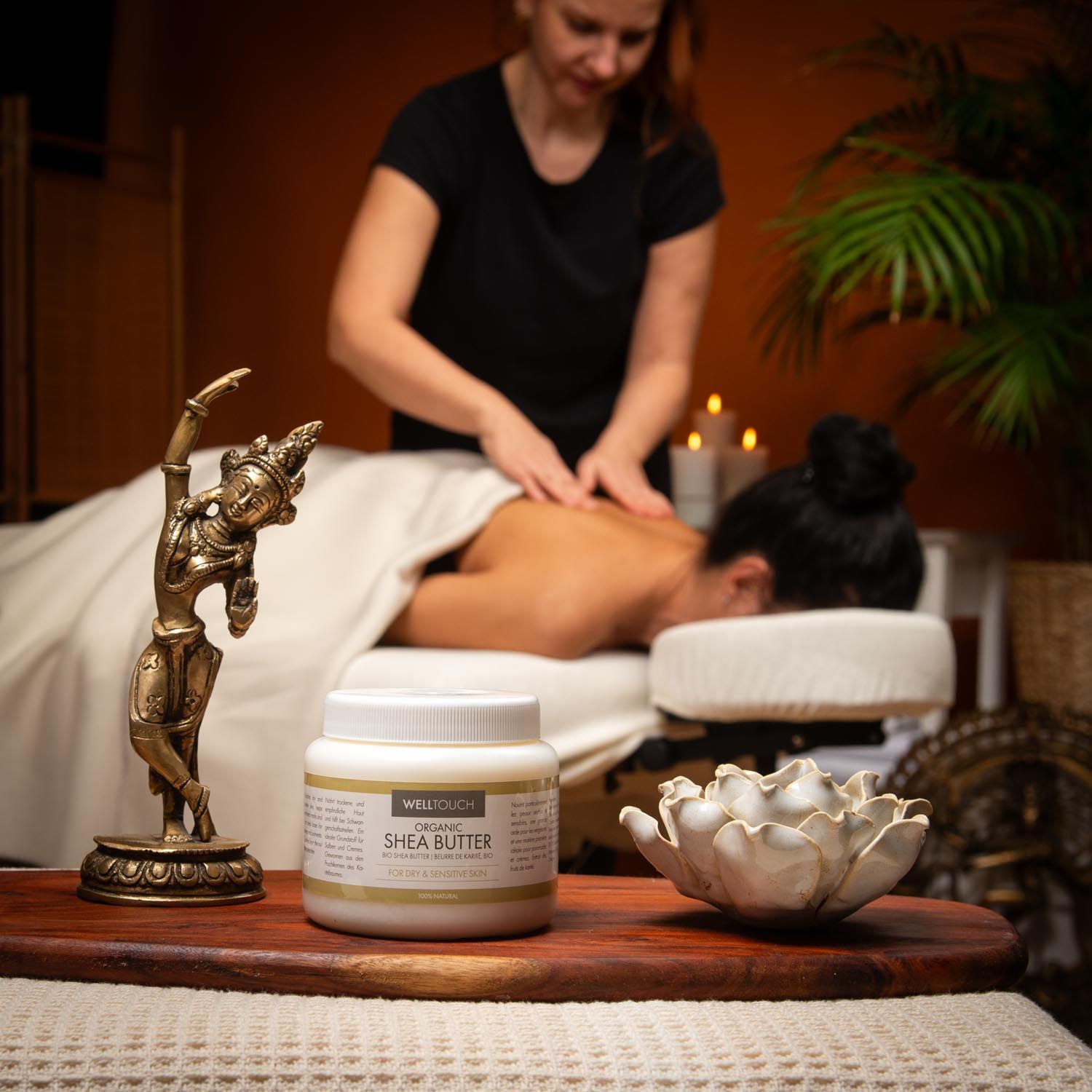 Witte pot Welltouch sheaboter op houten dienblad. Vrouw masseert persoon op massagetafel. Gouden beeldje en lotusbloem ernaast.