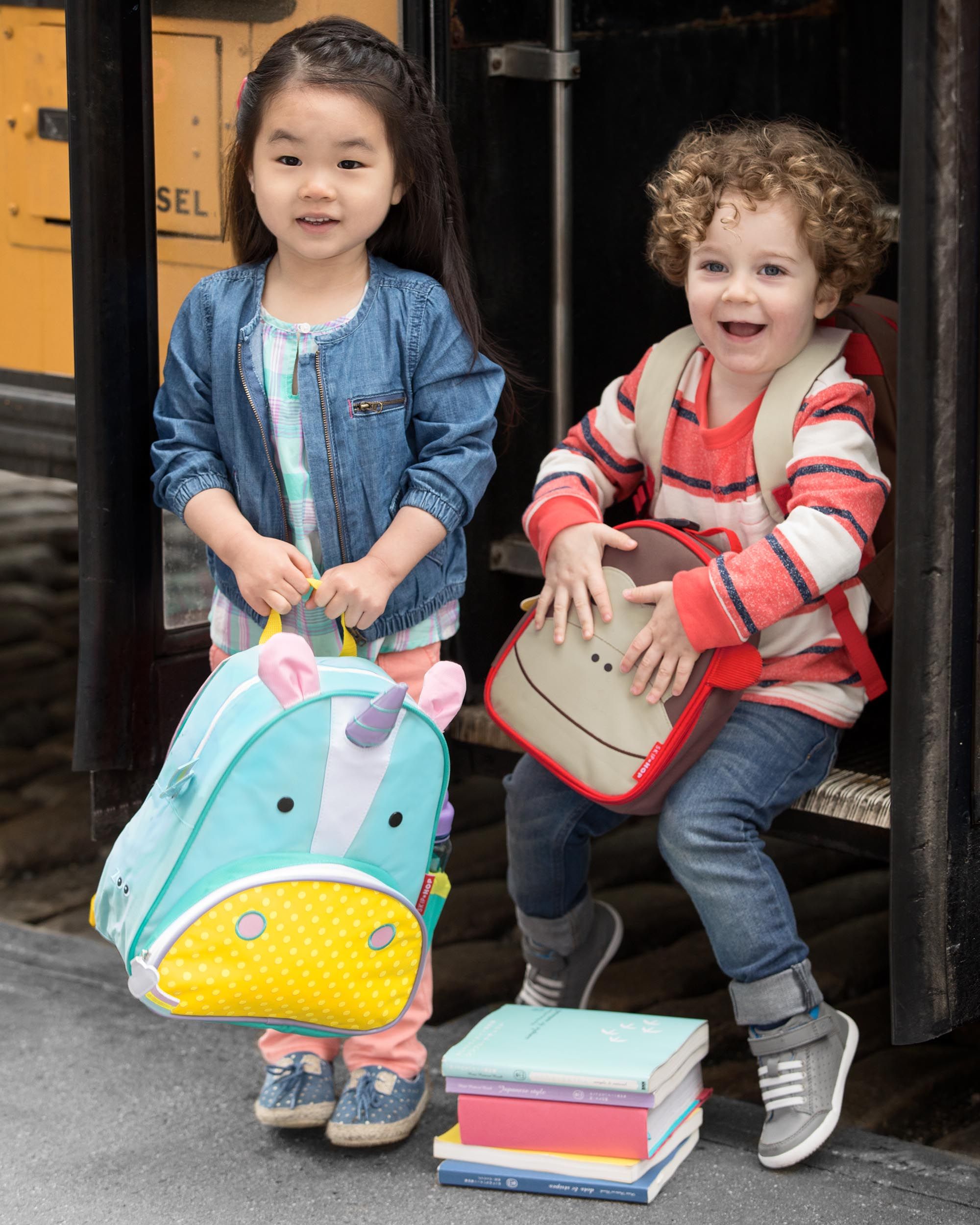 Twee kinderen staan bij een bushalte. Een met een eenhoornrugzak, de ander met een apenrugzak.