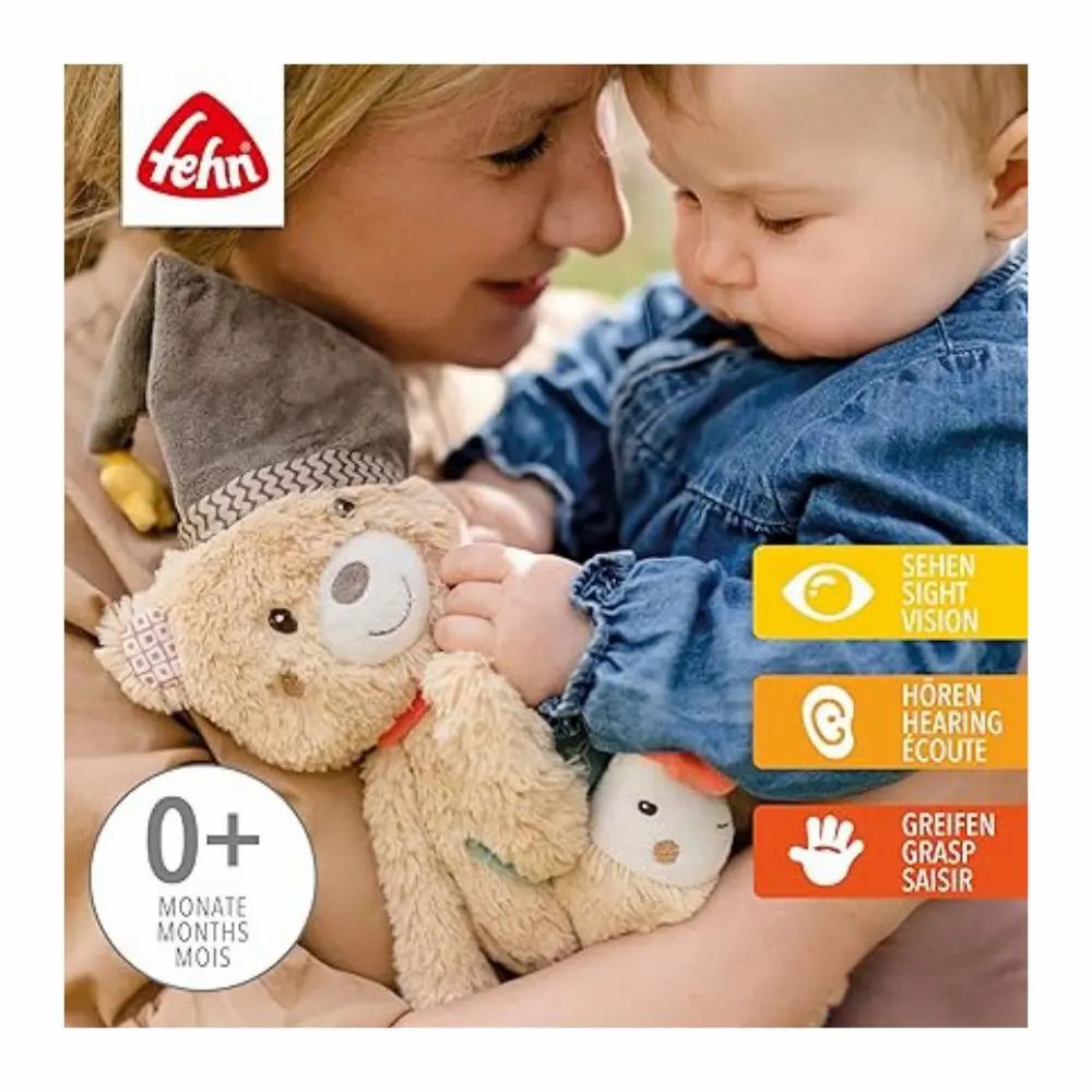 Bébé tient peluche avec bonnet, écharpe rouge, étoile et poisson. Marque: fehn. Avec texte et symboles.