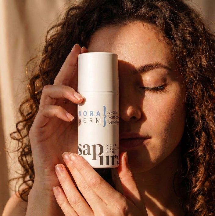 Vrouw houdt witte fles met pompdosering, opschrift 'NORADERM' en 'sapura', voor haar gezicht.