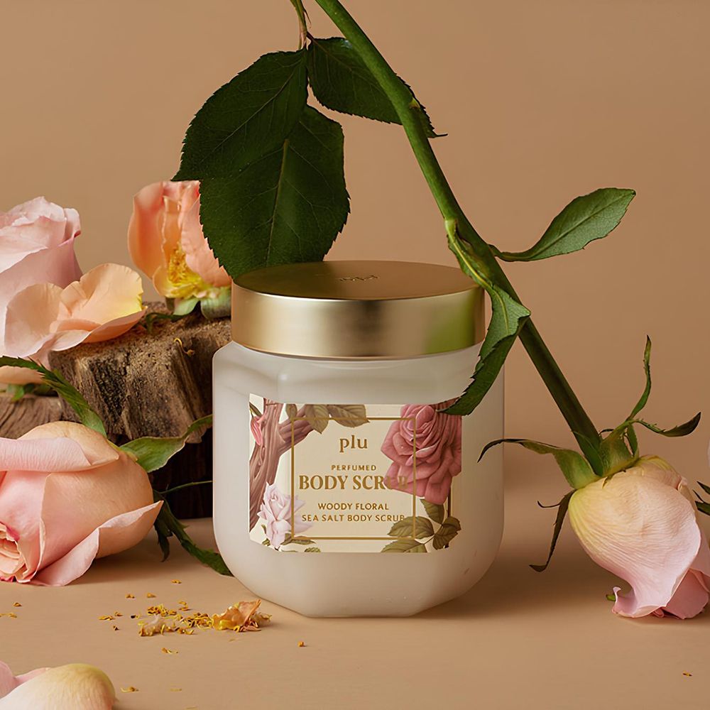 Pot en verre avec couvercle doré, entouré de roses. Étiquette avec nom du produit : PLU Perfumed Body Scrub.