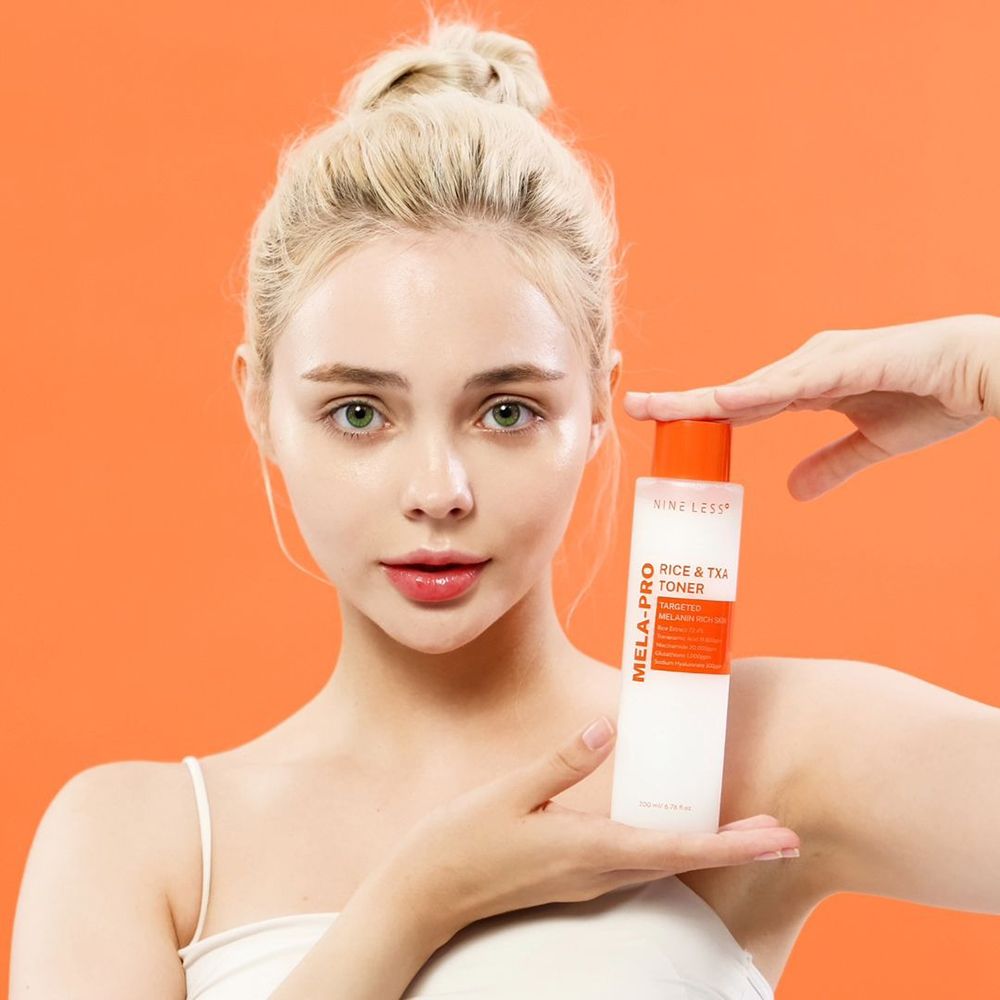 Een vrouw houdt een fles NINE LESS Mela-Pro Rice & TXA Toner vast. De fles is wit met oranje accenten. De achtergrond is oranje.