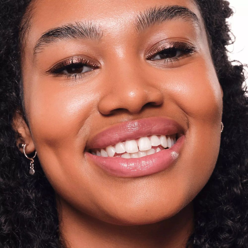 Femme avec eyeliner métallique or rose sur les paupières. Sourit à la caméra.