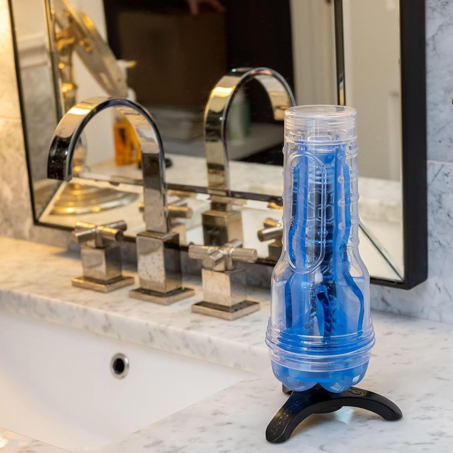 Een blauw, getextureerd product in een transparante houder met standaard op een marmeren oppervlak naast een kraan.