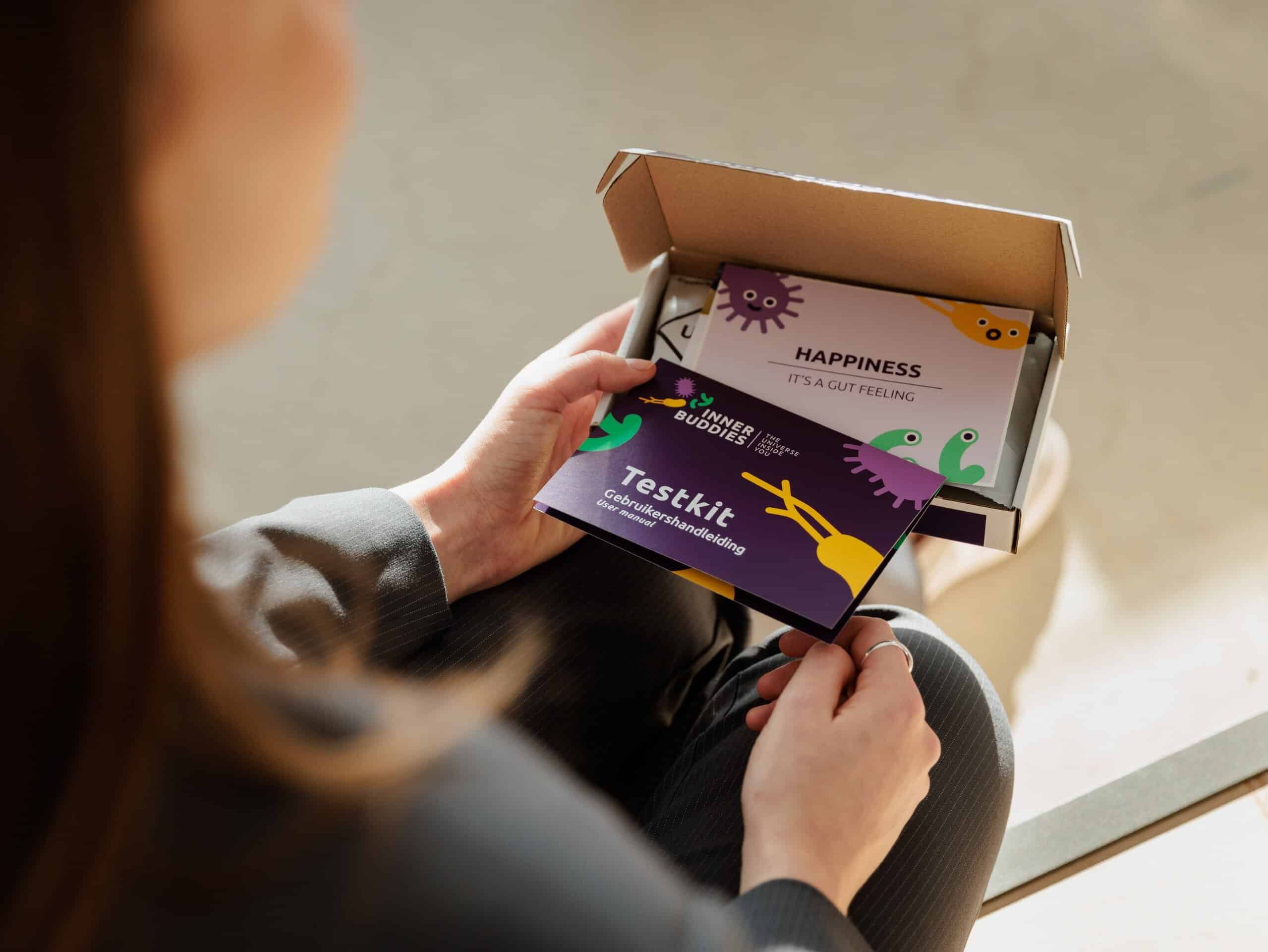 Een persoon houdt een 'Inner Buddies Testkit' brochure vast boven een geopende doos.