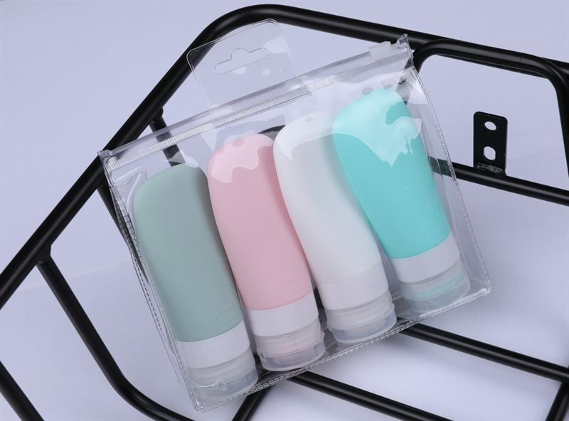 Quatre flacons de voyage colorés dans un sac transparent. Couleurs : vert, rose, blanc et turquoise. Sur un support noir.