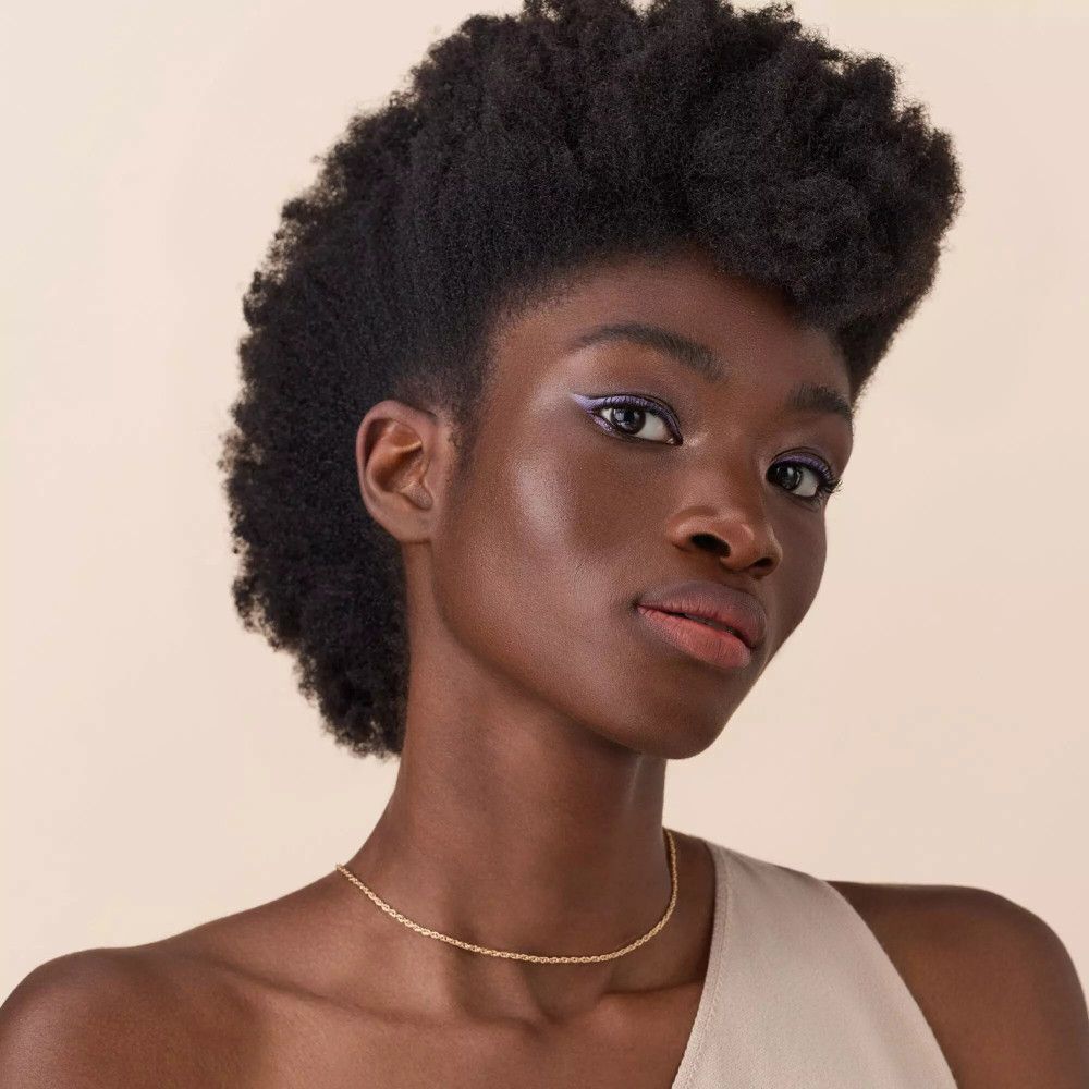 Portret van een vrouw met een donkere huid. Ze draagt een gouden ketting en paarse eyeliner.