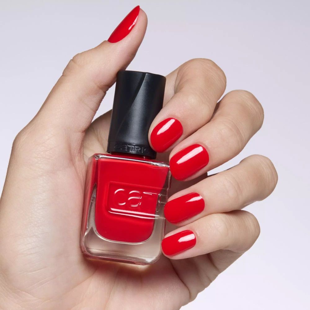 Main avec ongles rouges vernis et flacon de vernis. Marque Catrice sur le flacon.