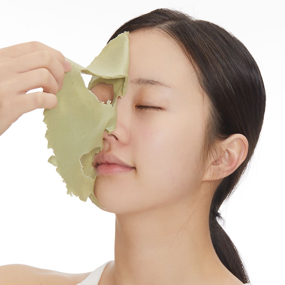 Visage de femme avec masque vert. Une main retire le masque. Fond blanc.