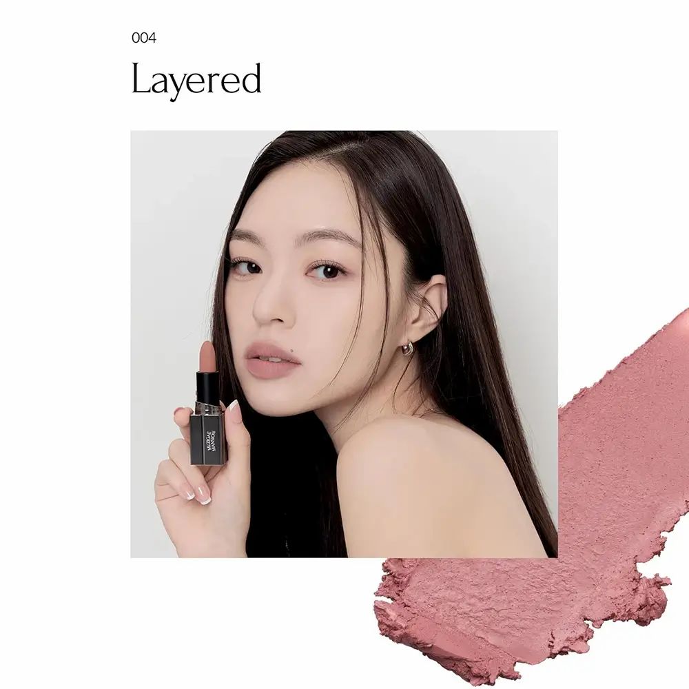 Vrouw houdt lipstick vast. Zwarte huls, zilverkleurige basis. MUZIGAE MANSION-logo. Tint 004 Layered.