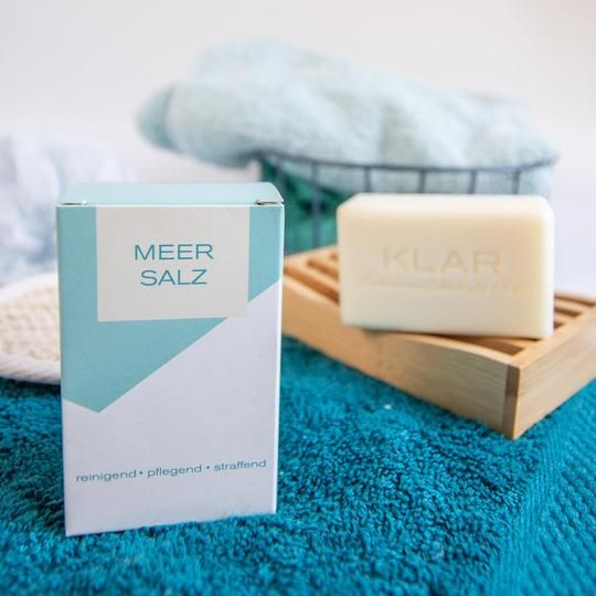 Klar-Seifen zeezoutzeep en verpakking op blauwe handdoek. Zeep op houten rekje. Verpakking: blauw-wit met "MEER SALZ".