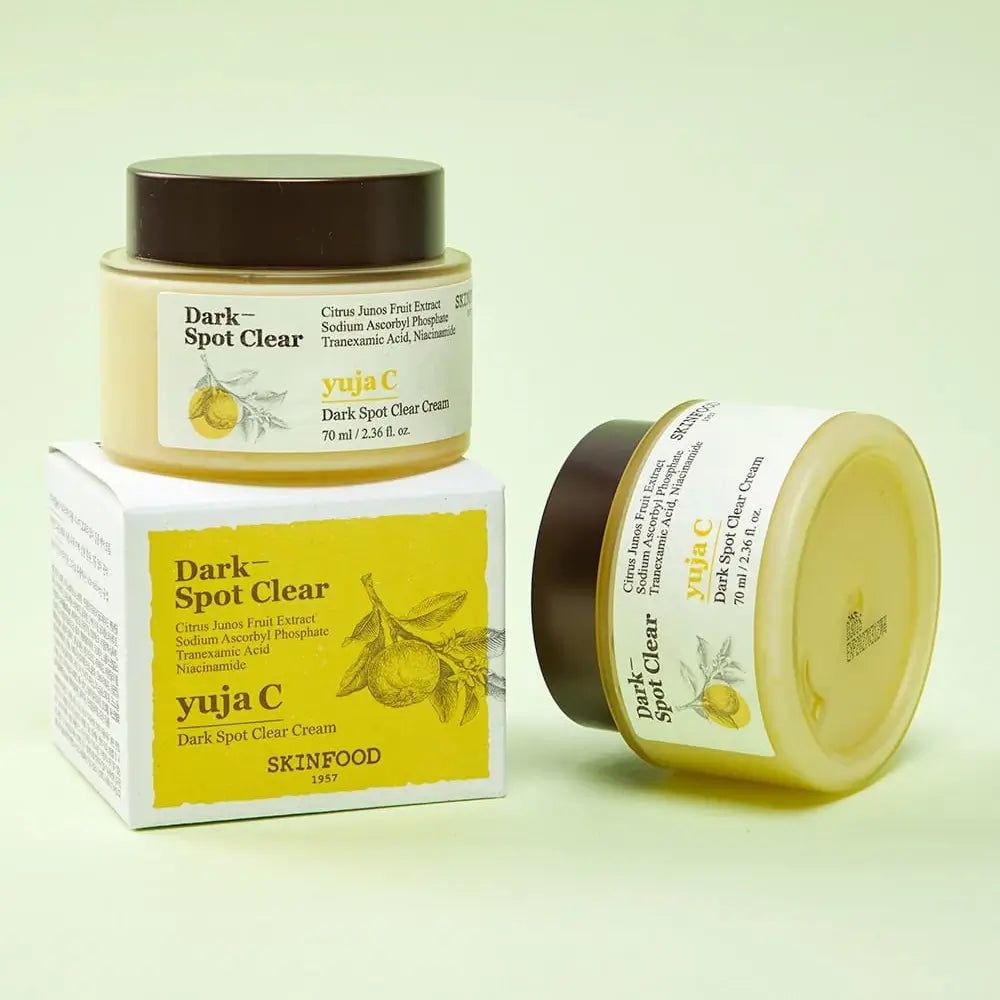 Twee crèmepotten en een doos. Opschrift: Dark Spot Clear, Yuja C, SKINFOOD. Gele crème in een pot met bruin deksel. Doos met productinformatie.